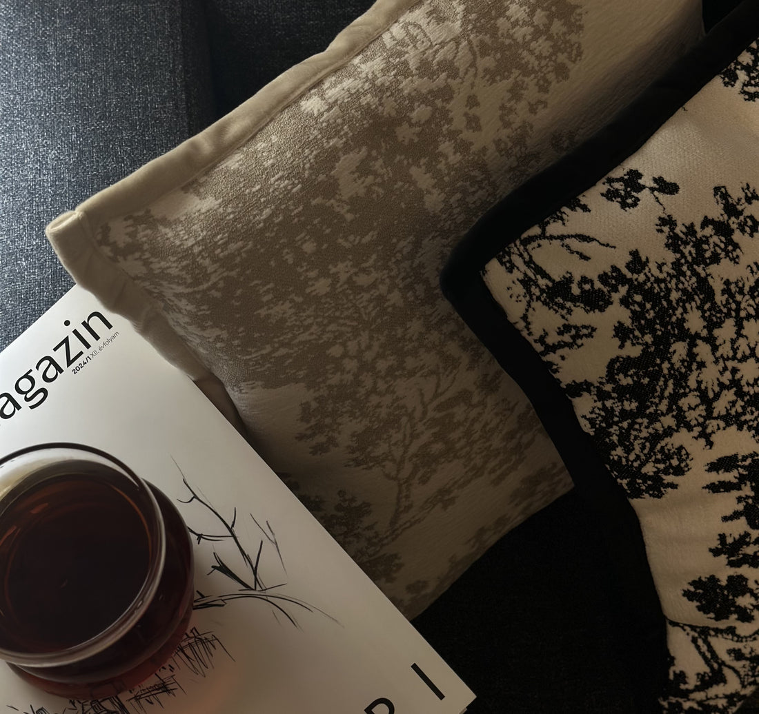 "Close-up of two stylish decorative pillows in neutral and black tones with intricate nature-inspired patterns, placed on a dark fabric sofa. A glass of tea rests on a magazine with a minimalist black-and-white cover."